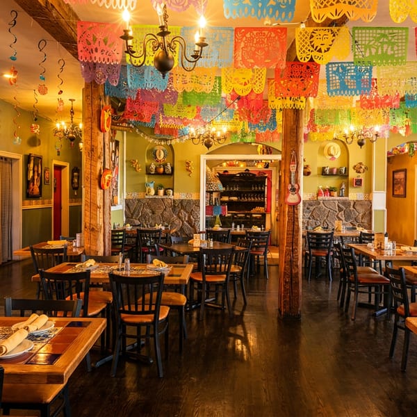 Vibrantly decorated Mexican restaurant dining room with colorful papel picado banners, wooden tables, and chairs.