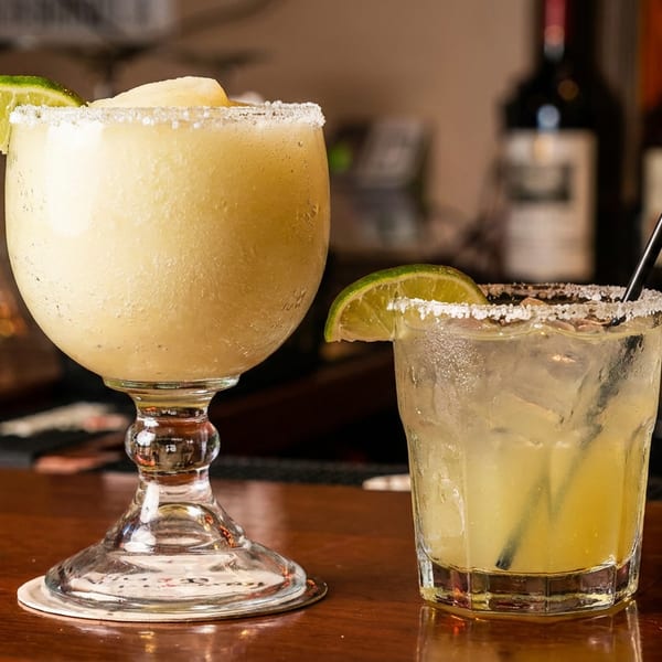 Two margaritas, one frozen and one on the rocks, garnished with lime and salt on a bar.
