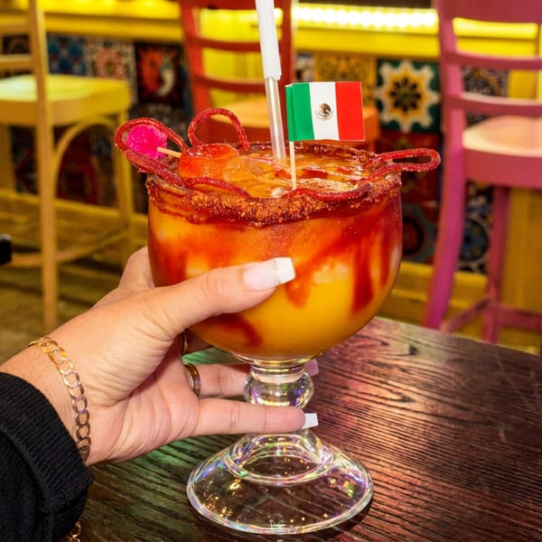 Hand holding a vibrant mango margarita with a Mexican flag, chili rim, and candy garnish.
