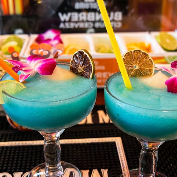 Two blue frozen cocktails garnished with lime, dried orange, and orchids on a bar.