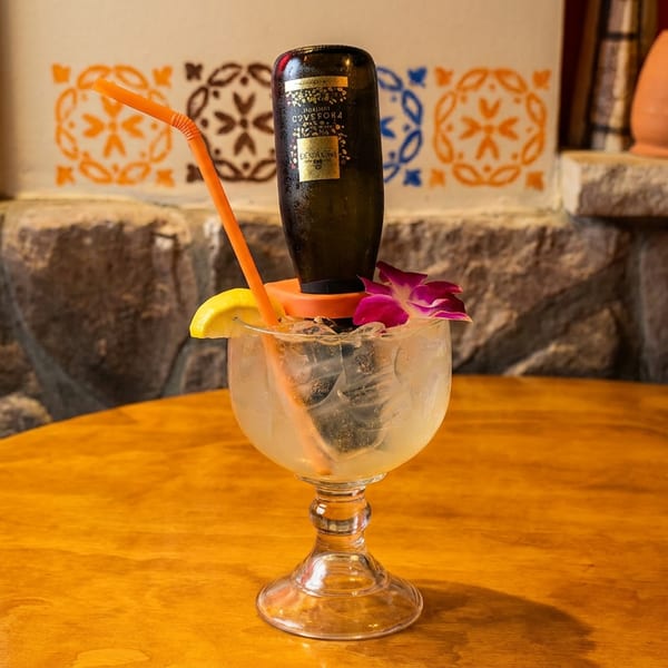 A large cocktail with a mini bottle of liquor inverted into it, garnished with a lemon and orchid.