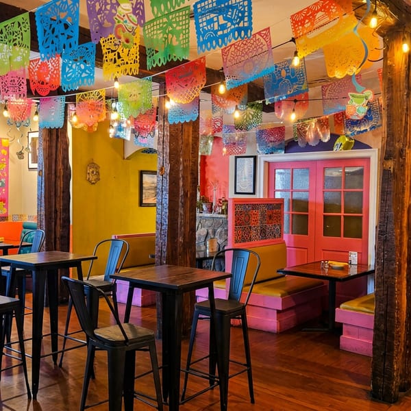Vibrant Mexican restaurant interior with colorful papel picado, wooden tables, and bright pink and yellow booths.