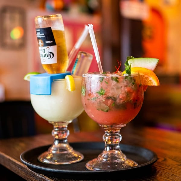 Two colorful cocktails, one with an inverted beer bottle, on a tray in a vibrant restaurant.