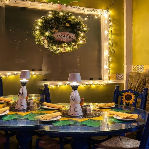 A festive restaurant interior with a blue table, sunflower chairs, and a lighted wreath.