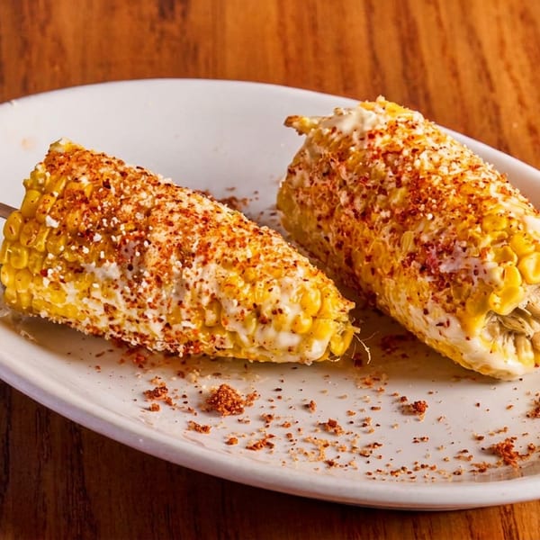 Two elotes, Mexican street corn, on a white plate with wooden skewers, topped with sauce and chili powder.