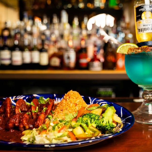 A blue margarita with a Corona bottle and a plate of shrimp, rice, and vegetables on a bar.