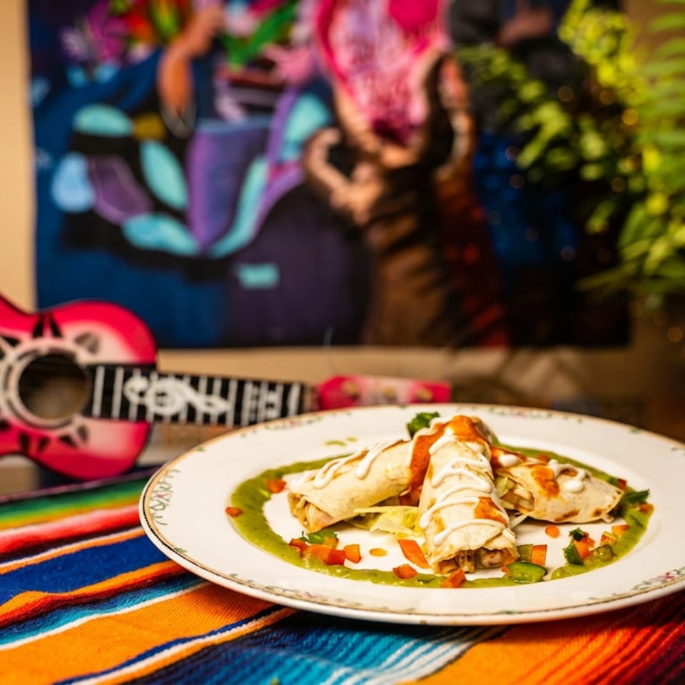 Three enchiladas with green and red sauce on a colorful striped blanket next to a pink toy guitar.